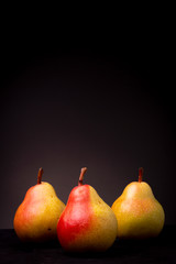 Vertical frame of three vibrant colourful yellow red Seckel pears on a black surface contrasted against a dark grey studio background