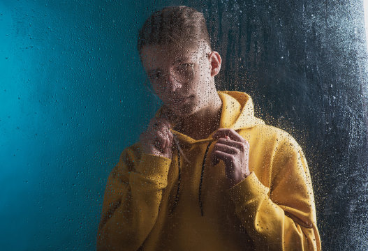 A young guy in a yellow hoodie is sad behind a blind window