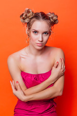 The girl in a red dress on a orange background in the studio. Blonde girl with two hair knots looking to the camera and smiling.