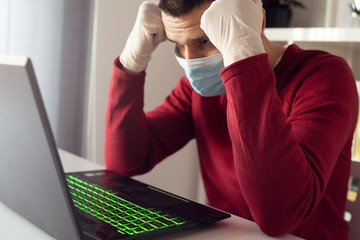 Young adult man caucasian wearing gloves on hands and protective mask on face while working from home office work by the table laptop preventing virus spread in epidemic quarantine hold head worried
