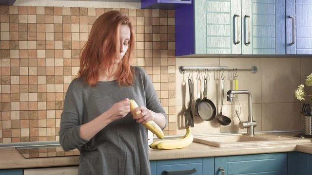 Attractive hungry redhead girl eats a big banana in the early morning on the weekend in the kitchen. Cleans the peel. Healthy eating, the right lifestyle. Sports breakfast