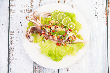 Spicy lemon squid with galangal, chilies, carrots, peppermint, spring onions and garlic on a plate on a white wooden floor.