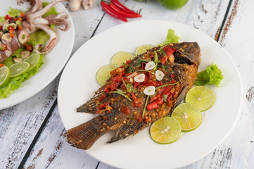 Fried tilapia with chili sauce,  lemon salad and garlic on a plate on a white wooden table.