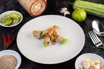 Spicy lime pork salad with bitter melon, galangal, chili, tomatoes and garlic in a white plate on a black cement floor.