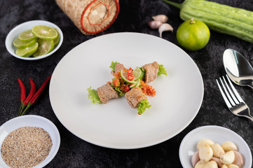 Spicy lime pork salad with bitter melon, galangal, chili, tomatoes and garlic in a white plate on a black cement floor.