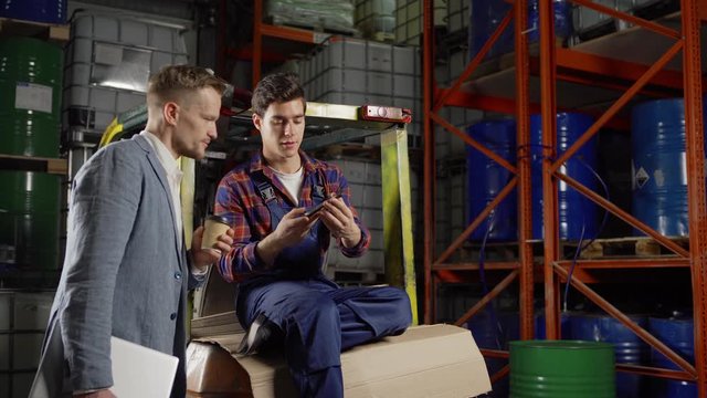 Tracking medium shot of smiling young forklift driver showing text message or picture on cell phone screen to middle aged manager drinking coffee during break in industrial storage