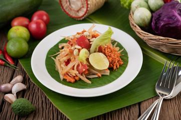 Thai papaya salad in a white plate on banana leaves with lime, tomatoes, eggplant, chili, garlic, peppers, salad, and peanuts.