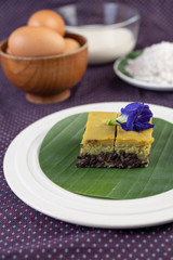 Black sticky rice and custard on a banana leaf in a white plate with butterfly pea flowers.