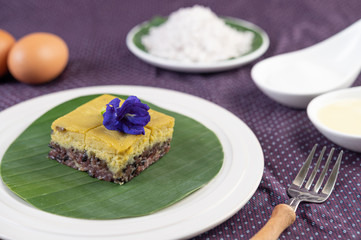 Black sticky rice and custard on a banana leaf in a white plate with butterfly pea flowers.
