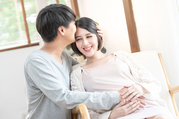 Happy excited pregnant couple having fun at home. Cheerful expectant mom sitting on couch dad kiss forehead, look out window smiling, laughing. Enjoying pregnancy mother’s day concept