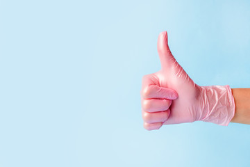 Closeup of female hand in pink cosmetic medical hygiene pharmacy gloves showing thumbs up sign against pastel blue background, copy space, minimal concept