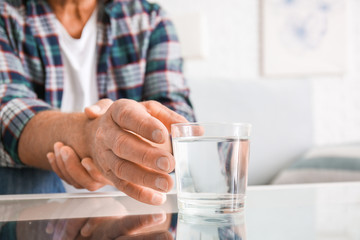 Senior man with Parkinson syndrome taking glass of water from table, closeup