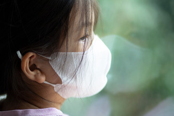 Asian 6 or 7 years old kid wearing medical mask.Little girl standing by the window and looking...
