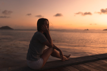 Fototapeta premium Beautiful girl at sunset on the sea in Thailand. Bright orange and yellow pink sky. Girl traveler enjoys the view. Woman in the tropics