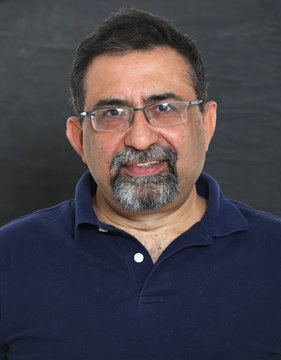 An Indian man with a goatee beard wearing spectacles against a dark background. Looking to camera