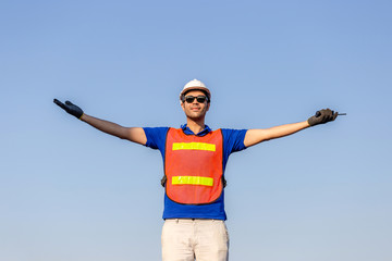 Successful young man with arms wide open, Happy engineer / foreman in hardhat and safety vest talks over blue sky background