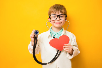 Cute child boy in doctor's uniform with stethoscope and heart in hand on yellow background. Copy...
