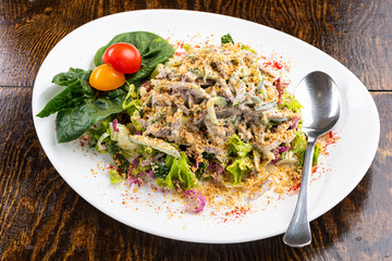 salad with meat, cheese, vegetables, mayonnaise, herbs on a white plate with a spoon on a wooden background