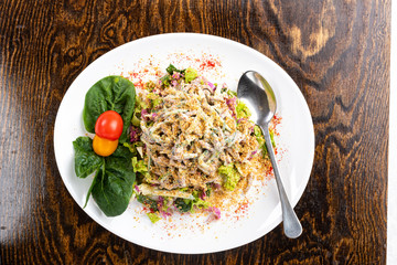salad with meat, cheese, vegetables, mayonnaise, herbs on a white plate with a spoon on a wooden background