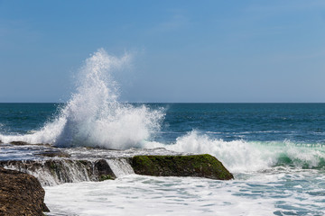 Fototapeta na wymiar Wave breaking on rock during low tide near Tanah Lot Temple, Bali, Indonesia. Deep blue ocean and sky in the background. 