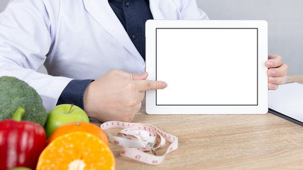 Nutritionist doctor hand pointing at digital tablet with blank screen for text, fresh fruits, vegetables, measuring tape and clipboard on wooden table. Healthy diet, nutrition and weight loss concept.