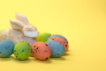 Colorful easter eggs painted in pastel colors and white rabbit hare toy with virus mask on yellow background. Easter greeting card, Corona virus (COVID19) protection concepts