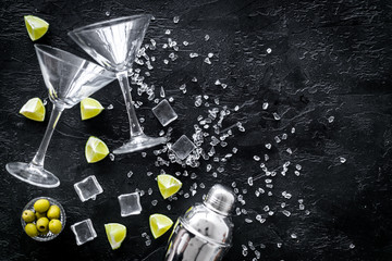 Bar background with martini glasses, shaker, ice, lemon and olives on black table top-down copy space