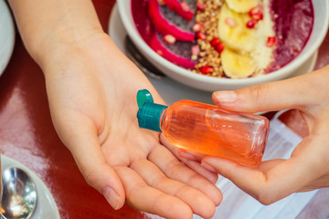 woman useing antibacterial gel on hands before start to eating her healthy food in tropical asian cafe . smoothie bowl , raw cake and coffee on table with sanitizer on bottle