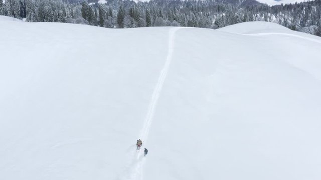 Aerial view man on snowmobile riding snowboarder on snowy slope in winter resort. People riding on snowmobile and snowboarders on snowy highlands in abonded winter village. Follow view from drone.