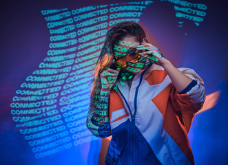 Serious and attractive female standing in neon studio with text projection, wearing bright blazer and holding her holographic sunglasses. Cyberpunk concept