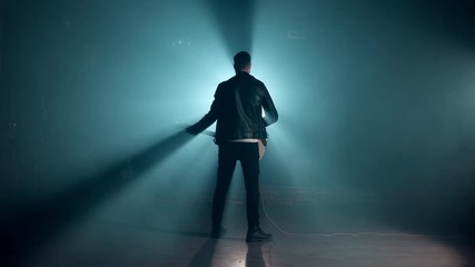 Rock guitarist man standing his back on smoky stage and masterfully playing electric guitar. View of musician raises hand up at end as gesture and symbol of success, freedom and victory