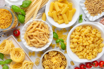 Variety of types and shapes of dry pasta - fusilli, penne, fettuccine nests, mafaldine, shells, bowtie and wheels - in white bowls 