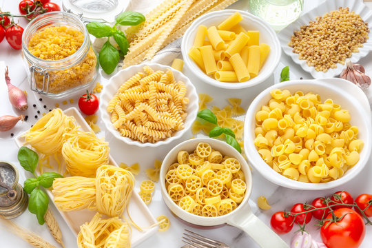 Variety of types and shapes of dry pasta - fusilli, penne, fettuccine nests, mafaldine, shells, bowtie and wheels - in white bowls 