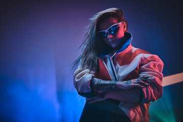 Stylish and futuristic young model posing in vivid neon studio lights of neon text projection over pink background, wearing racing blazer and holographic sunglasses. Cyberpunk concept