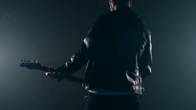 Rock guitarist man in leather jacket standing his back in smoky studio or stage masterfully playing electric guitar. View of unrecognizable musician in the spotlight. Slow motion 4K.