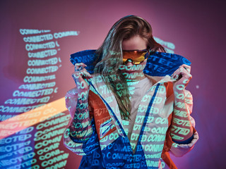 Attractive and futuristic young model posing in smooth neon studio lights of text projection over pink background, while holding the collar of her racing blazer and wearing holographic sunglasses