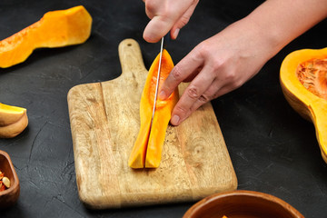 Butternut squash pieces on wooden chopping board on black background. Female hands with steel knife, closeup. Woman cuts a pumpkin on stone table. Cooking sweet winter squash