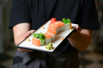 Waiter holds a plate of tasty food, sushi rolls. Japanese cuisine in restaurant. Waiter in a black uniform serving food.