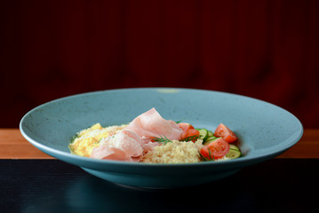 Mix of bulgur, ham prosciutto with sauce and vegetable salad - tomatoes and cucumbres served on a blue plate.