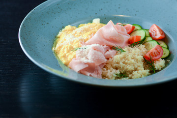 Mix of bulgur, ham prosciutto with sauce and vegetable salad - tomatoes and cucumbres served on a blue plate.