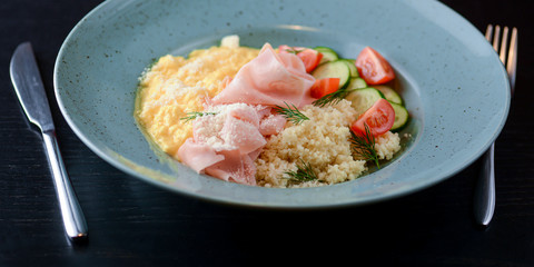 Mix of bulgur, ham prosciutto with sauce and vegetable salad - tomatoes and cucumbres served on a blue plate.
