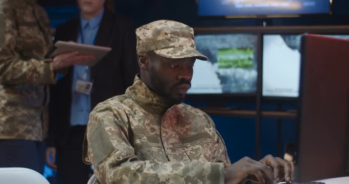Close up of African American young man in camouflage uniform and cap sitting at laptop computer and typing on keyboard in monitoring room in army. Soldier working in military center.