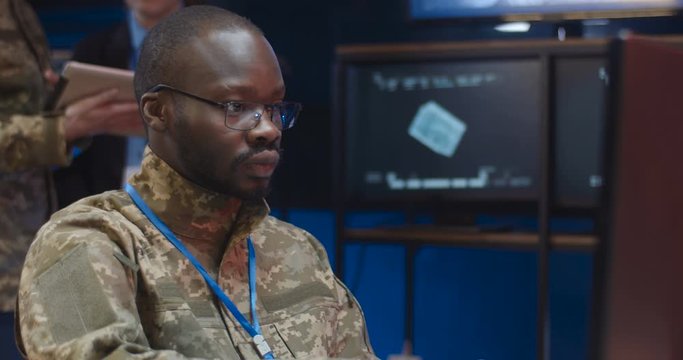 Close up of African American young man in camouflage clothes and glasses sitting at laptop computer and typing on keyboard in monitoring room in army. Soldier working in military data center.