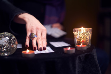 Close-up of female fortuneteller's hand with fortune-telling ring on cards at table with candles in dark room, blurred background