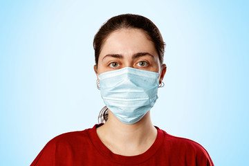 Portrait of a brunette woman in a medical mask and a red t-shirt, on a light blue background. Copy space. Concept of pandemic and protection against viral infection