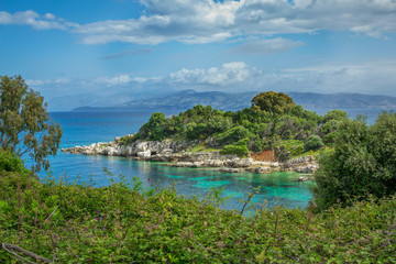 Fototapeta na wymiar Beautiful summer landscape of sea lagoon with turquoise calm water, cliffs and rocks on the shore, meadow with green grass, trees and bushes, mountains on the horizon. Corfu Island, Greece