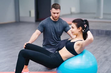 Tuinposter Trainer helping client workout on exercise ball at the gym. Sport and assistance concept. Doing exercises on fitball © Vadim