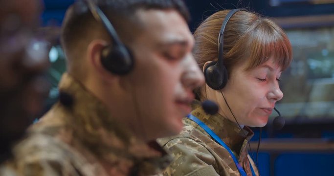 Close up of Caucasian male officer in headset working as dispatcher in monitoring room. Soldier answering calls on hot line of army controlling center. Multiethnic male and female co-workers beside.