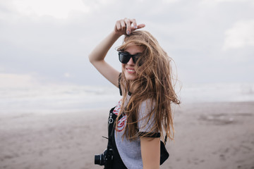 Glamorous female photographer smiling on sky background in windy day. Outdoor shot of stylish funny girl expressing happiness while posing at beach with camera.