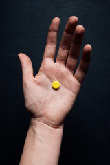 One hand holds one small yellow pill on a dark background. View from above. The medicine.